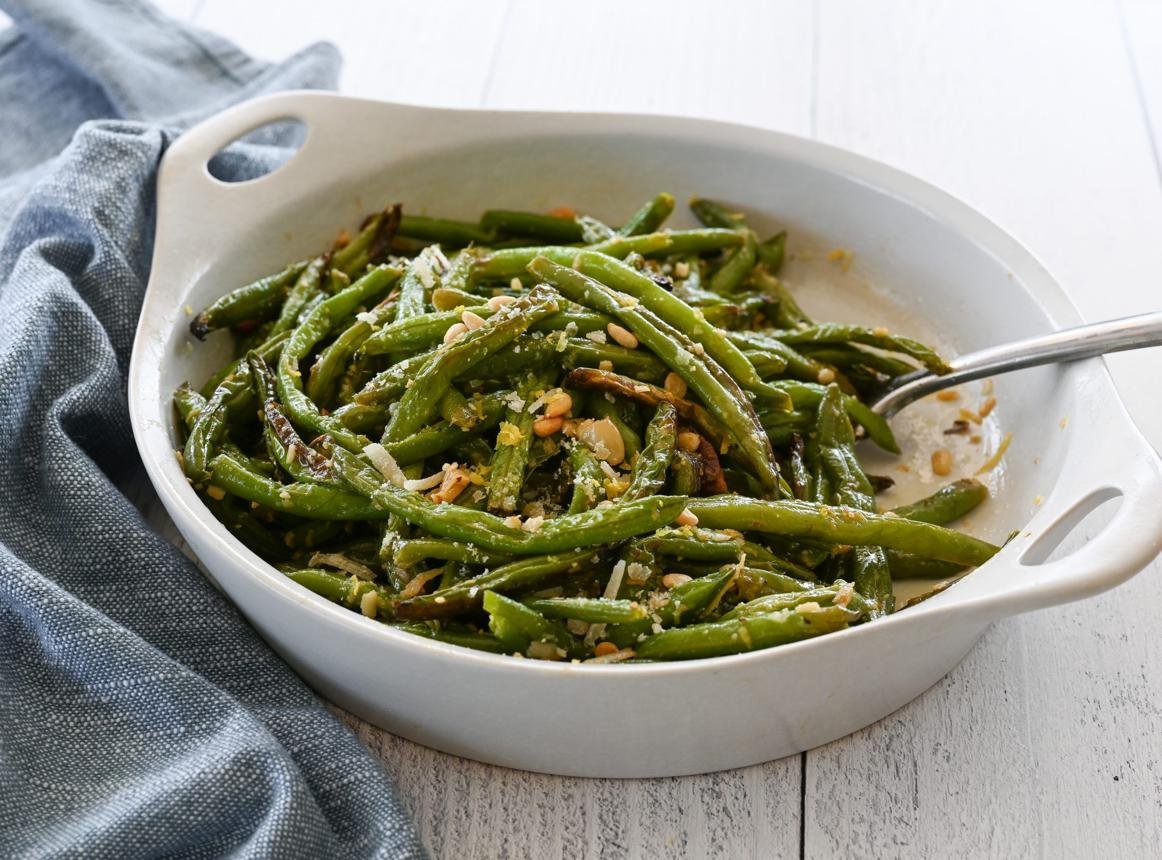 Sautéed green beans with slivered almonds in a white baking dish on a pale wood table.