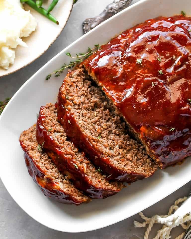 Slices of glazed meatloaf on a white platter, with mashed potatoes and thyme nearby.