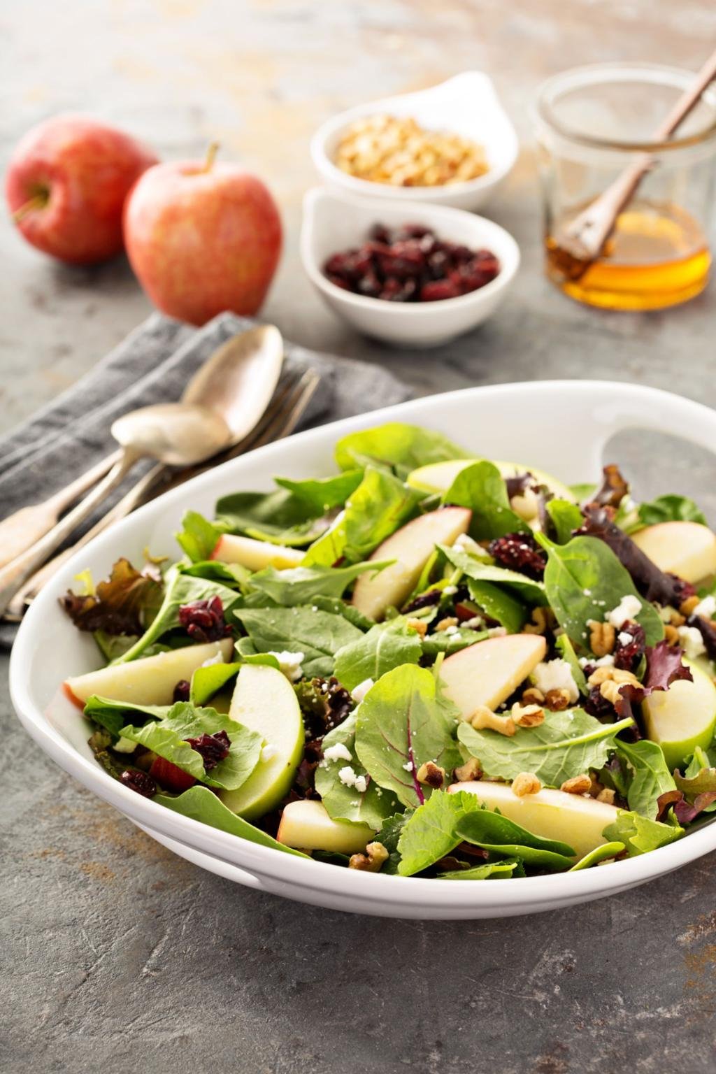 Bright salad bowl with mixed greens, apple slices, crumbled cheese, and walnuts on a gray surface with apples and jars in the background, informative image of a fruit and greens salad.