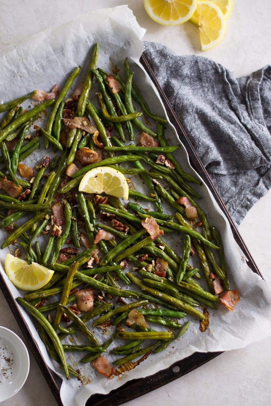 Sheet pan roasted green beans with bacon bits and lemon wedges, on parchment paper with a gray cloth nearby