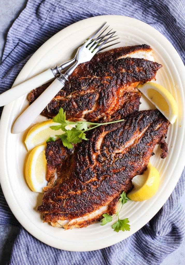 Two blackened fish fillets on a white plate with lemon wedges and parsley, ready to serve.