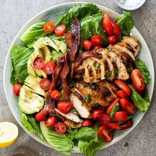 Grilled chicken slices on a bed of romaine with avocado, cherry tomatoes, and greens on a round plate, lemon wedge nearby