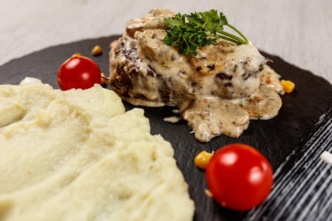 Creamy mushroom-sauce chicken fillet garnished with parsley, served with mashed potatoes and cherry tomatoes on a dark slate plate.