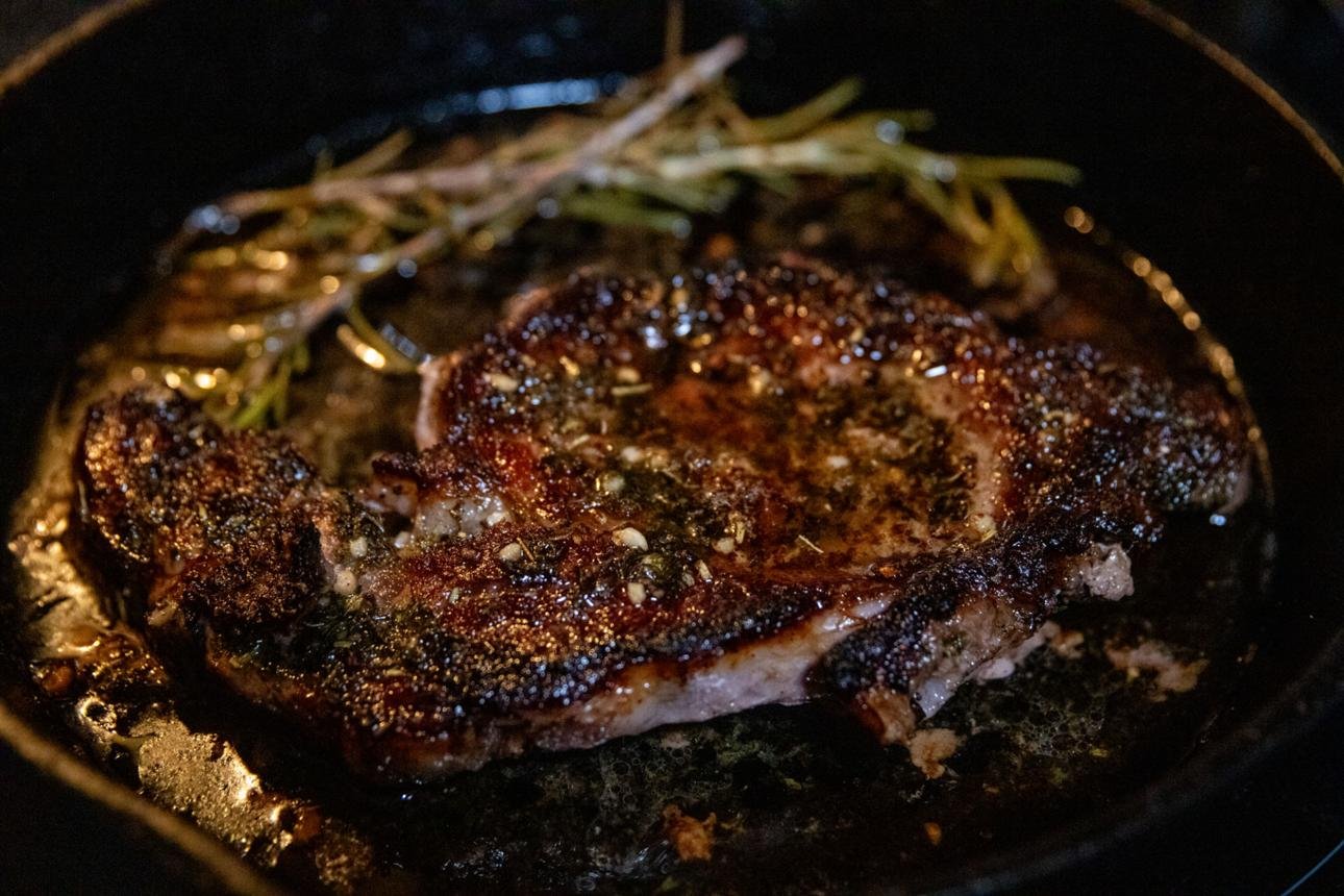 Thick steak with a dark, caramelized crust cooking in a skillet with herbs nearby.