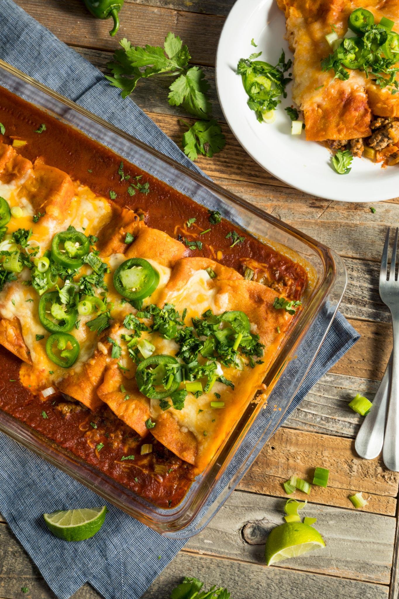 Baked cheese enchiladas topped with jalapeño slices and cilantro in red sauce in a glass baking dish on a wooden table, with a plate of enchiladas nearby and lime wedges nearby.