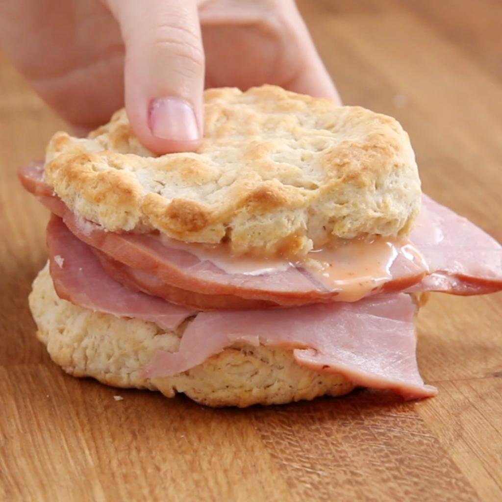 Hand placing the top half of a ham biscuit sandwich on a wooden board.
