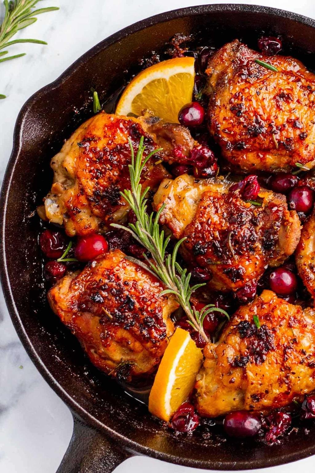 Cast iron skillet with roasted chicken pieces, orange slices, rosemary, and cranberries; glossy glaze and herbs brighten the dish