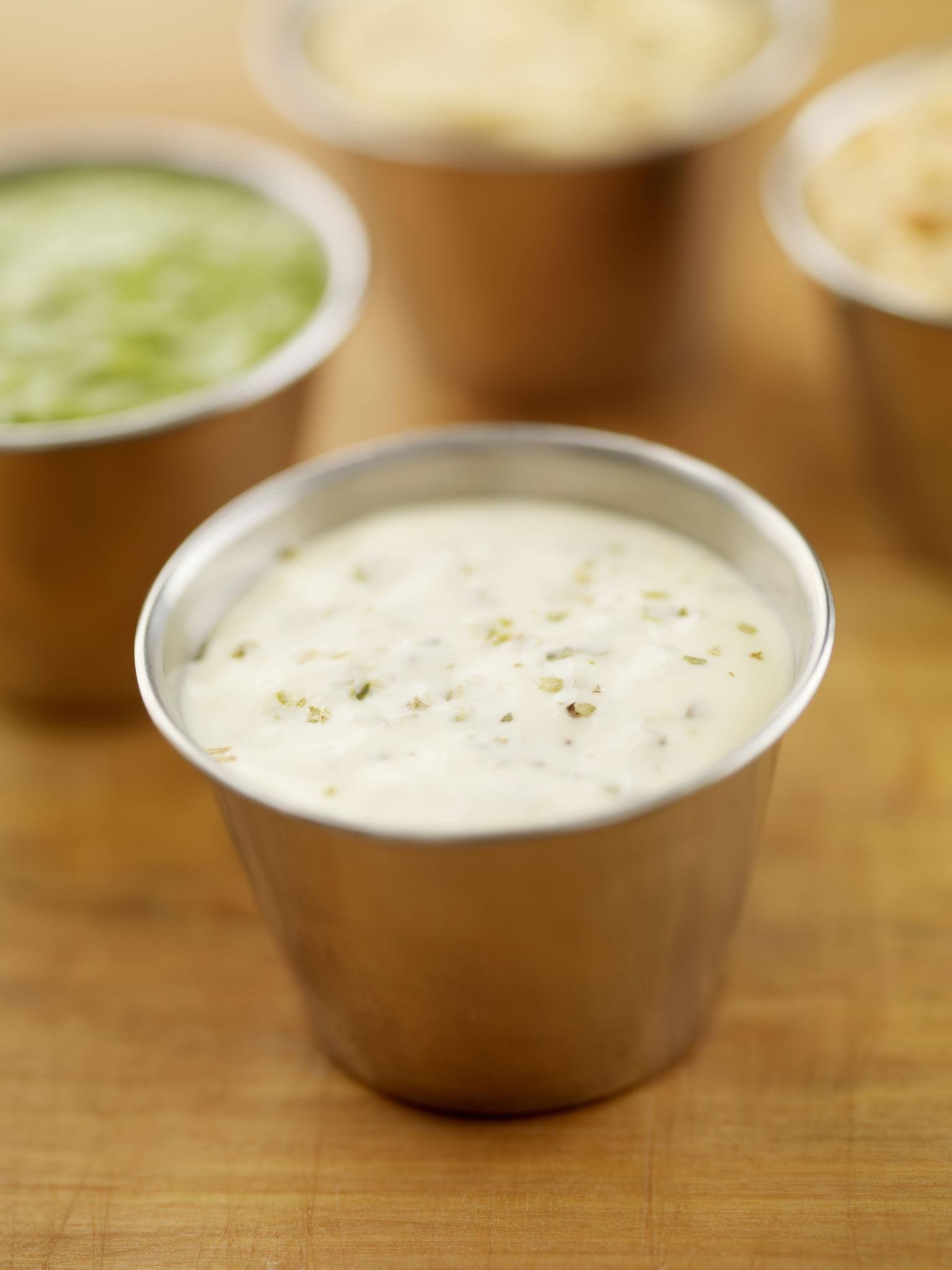 Cup of ranch dressing with herbs on a wooden table, with blurred green and beige dips in the background.