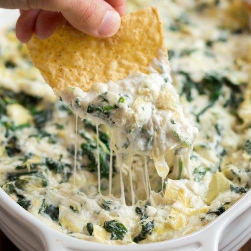 Close-up of a tortilla chip being dipped into warm spinach and artichoke dip with melted cheese strings flowing from the chip.