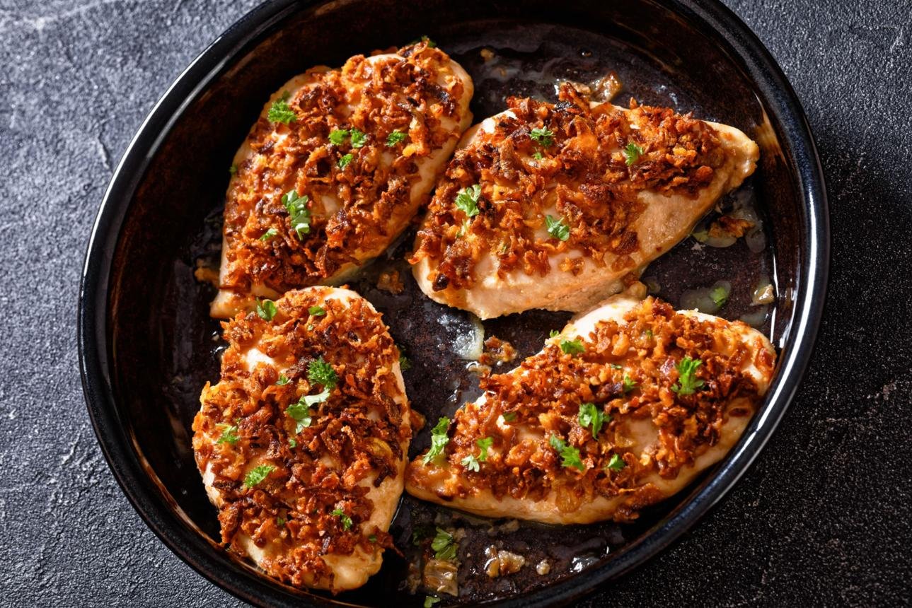 Four chicken breasts baked with crispy fried onions and parsley in a dark roasting pan.