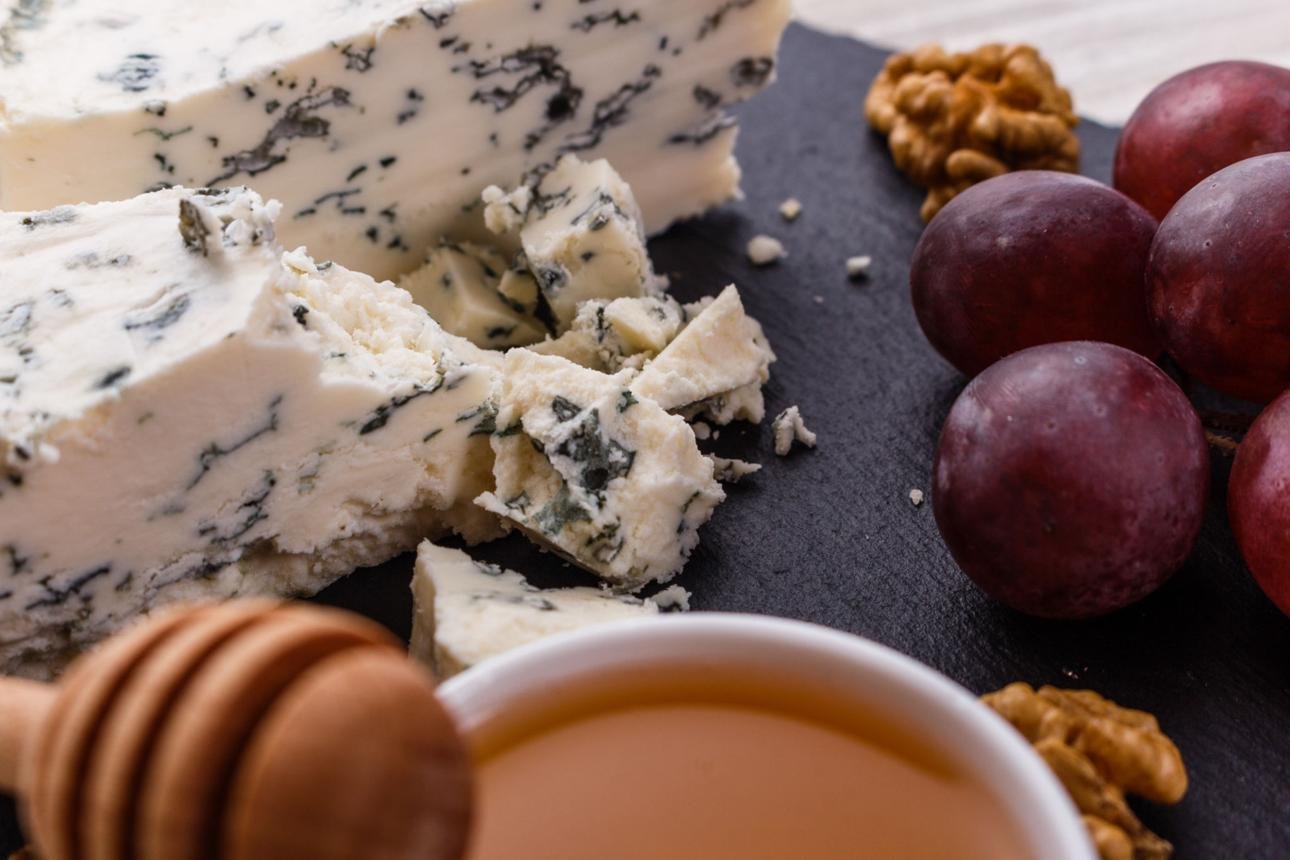 Blue cheese wedges with blue veins broken into crumbles on a dark slate board, surrounded by red grapes, walnuts, and a honey dipper with honey.