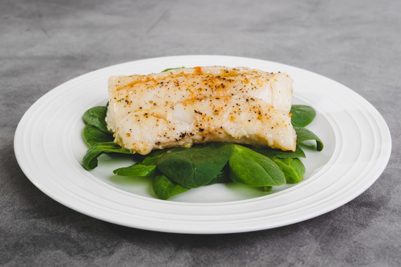 Piece of seasoned white fish resting on a bed of fresh spinach on a white plate.