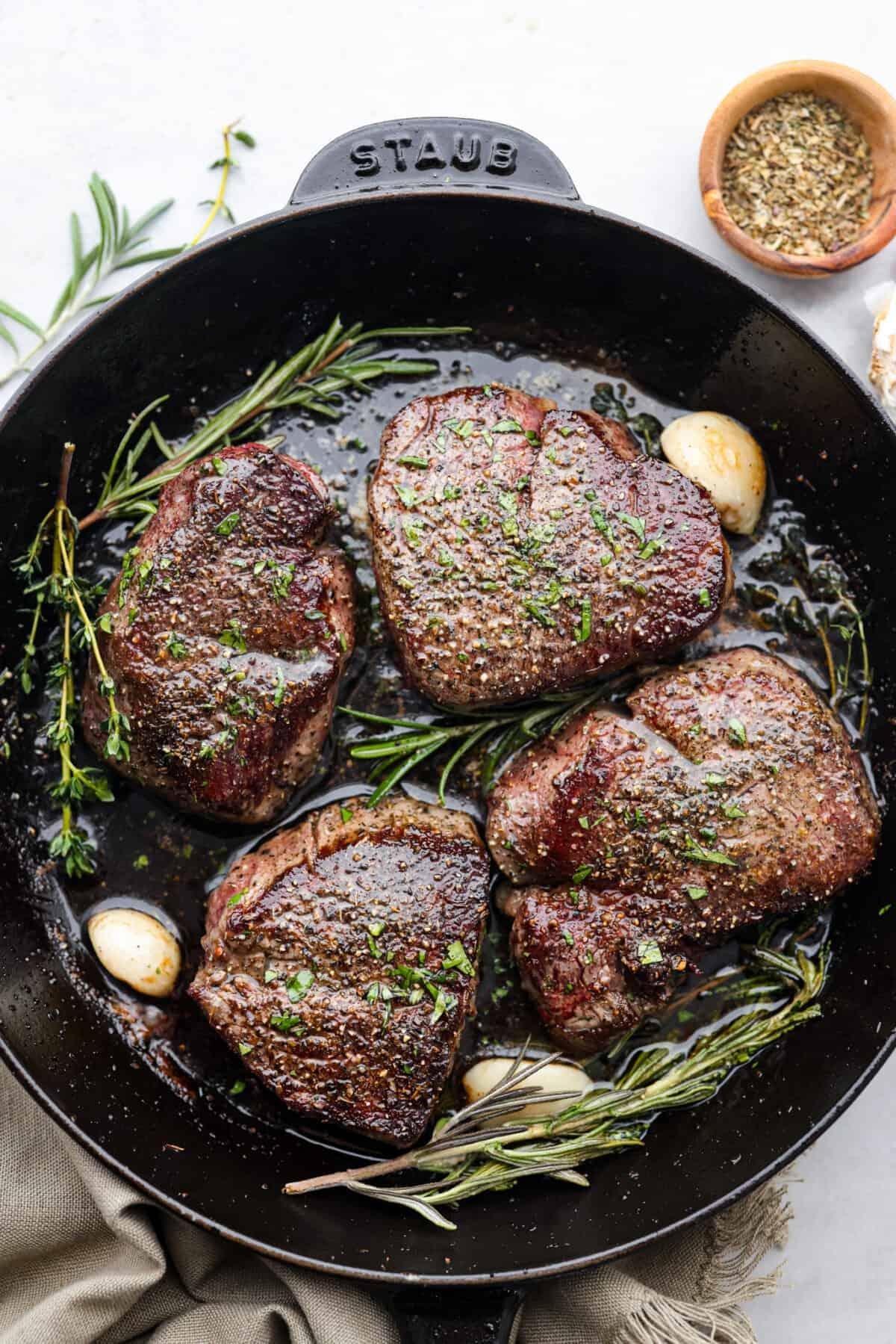 Five beef steaks seared with herbs (rosemary, thyme) in a black cast-iron Staub pan with garlic cloves nearby.