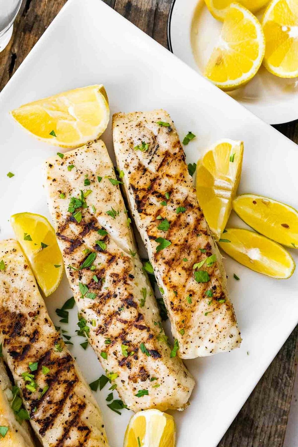 Grilled white fish fillets on a white plate, sprinkled with parsley and served with lemon wedges.