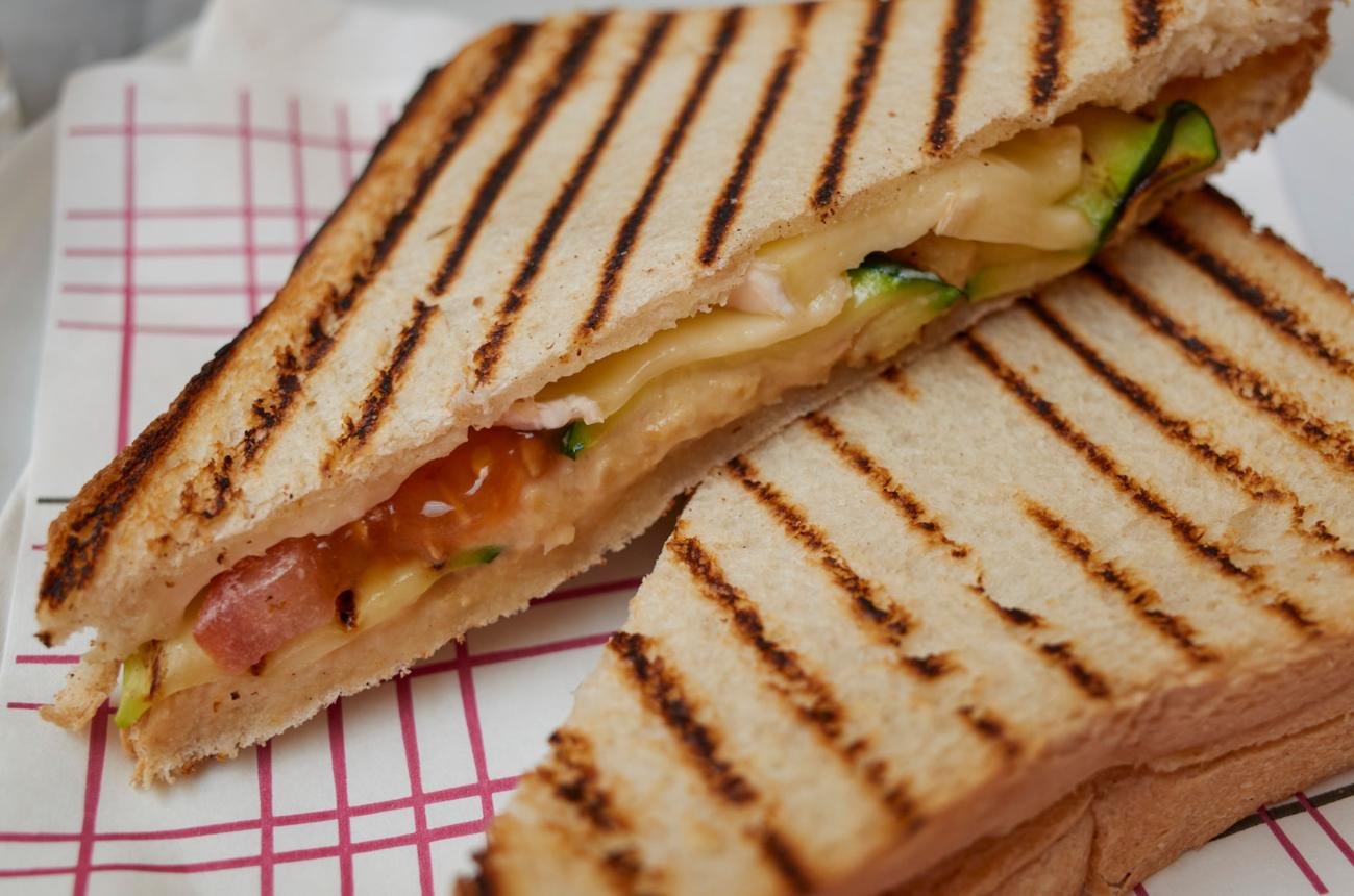 Grilled ham and cheese panini with cucumbers, cut into two diagonal halves with charred grill lines on white bread.