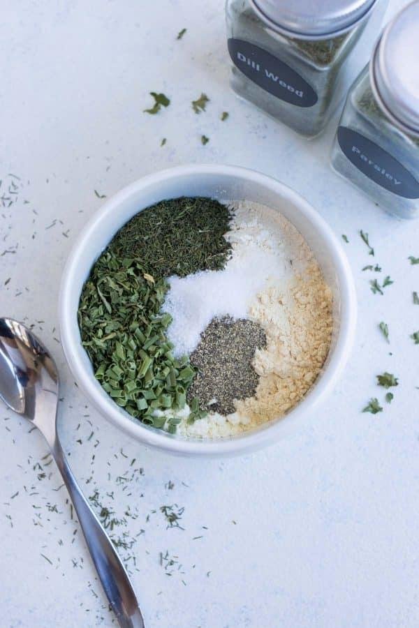 Top‑down view of a white bowl with a divided herb and spice blend (dill weed, dried greens, salt, pepper, and powders); a spoon and two labeled jars nearby.