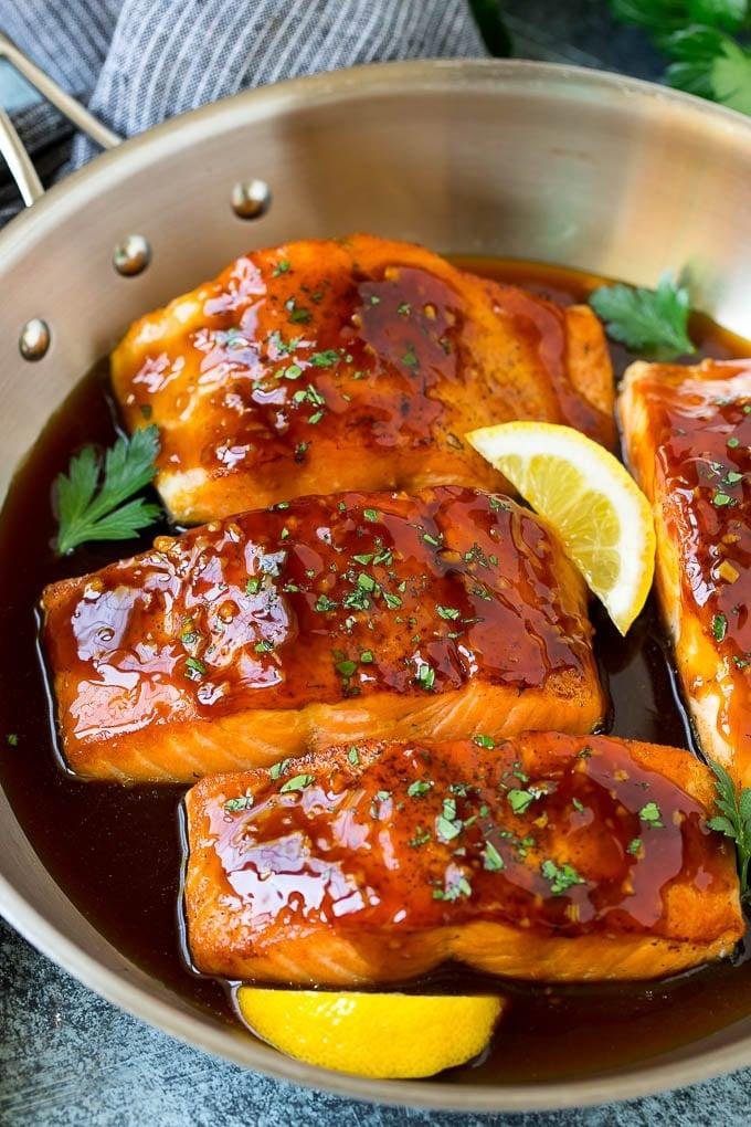 Salmon fillets with a glossy brown glaze cooking in a skillet, garnished with parsley and lemon wedges