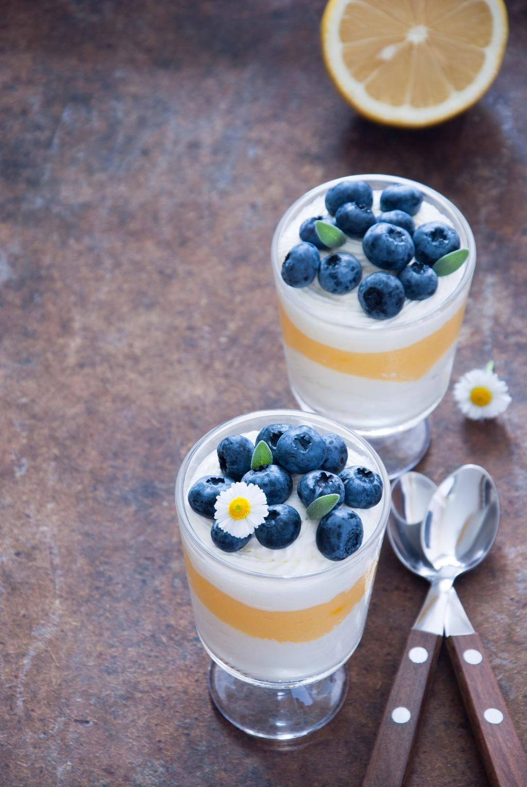Two dessert parfaits in stem glasses with blueberries, lemon curd layers, and cream on a rustic table.