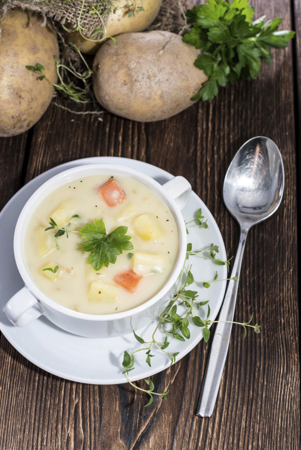 Creamy potato soup in a white cup with potato chunks and parsley, on a white saucer over a dark wooden table, with a spoon nearby.