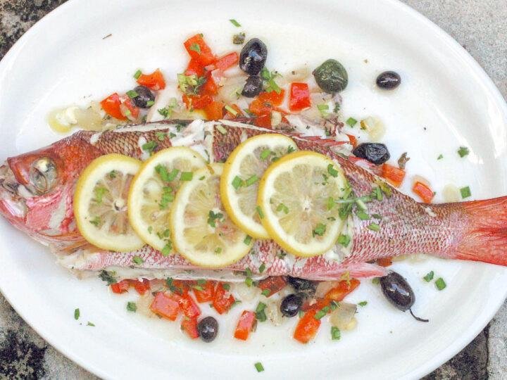 Whole red snapper on a white plate, garnished with lemon slices, herbs, and a diced pepper relish with olives around it
