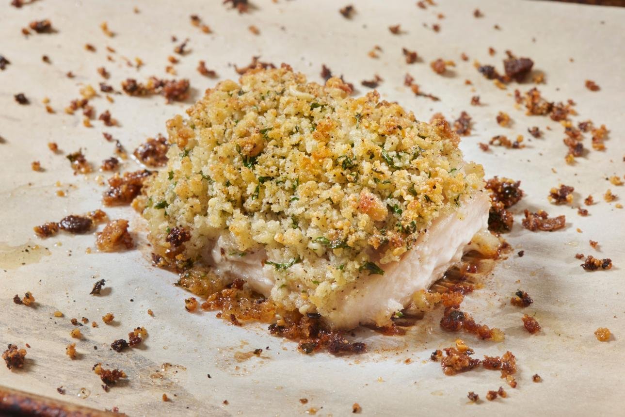 Herb-crusted baked fish fillet with a golden breadcrumb topping on parchment, crumbs scattered around.