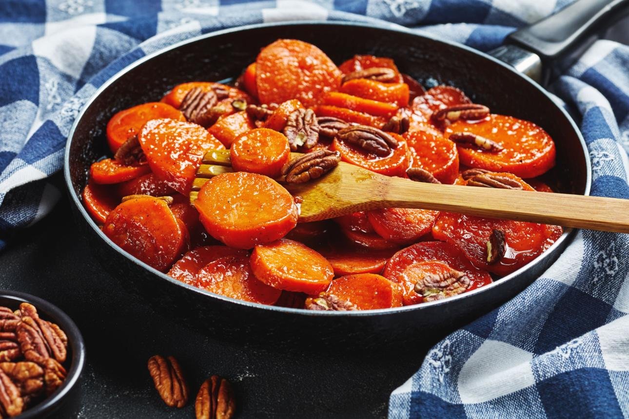 Skillet with glazed carrot coins and pecans, a wooden spoon resting on top on a blue checkered cloth background