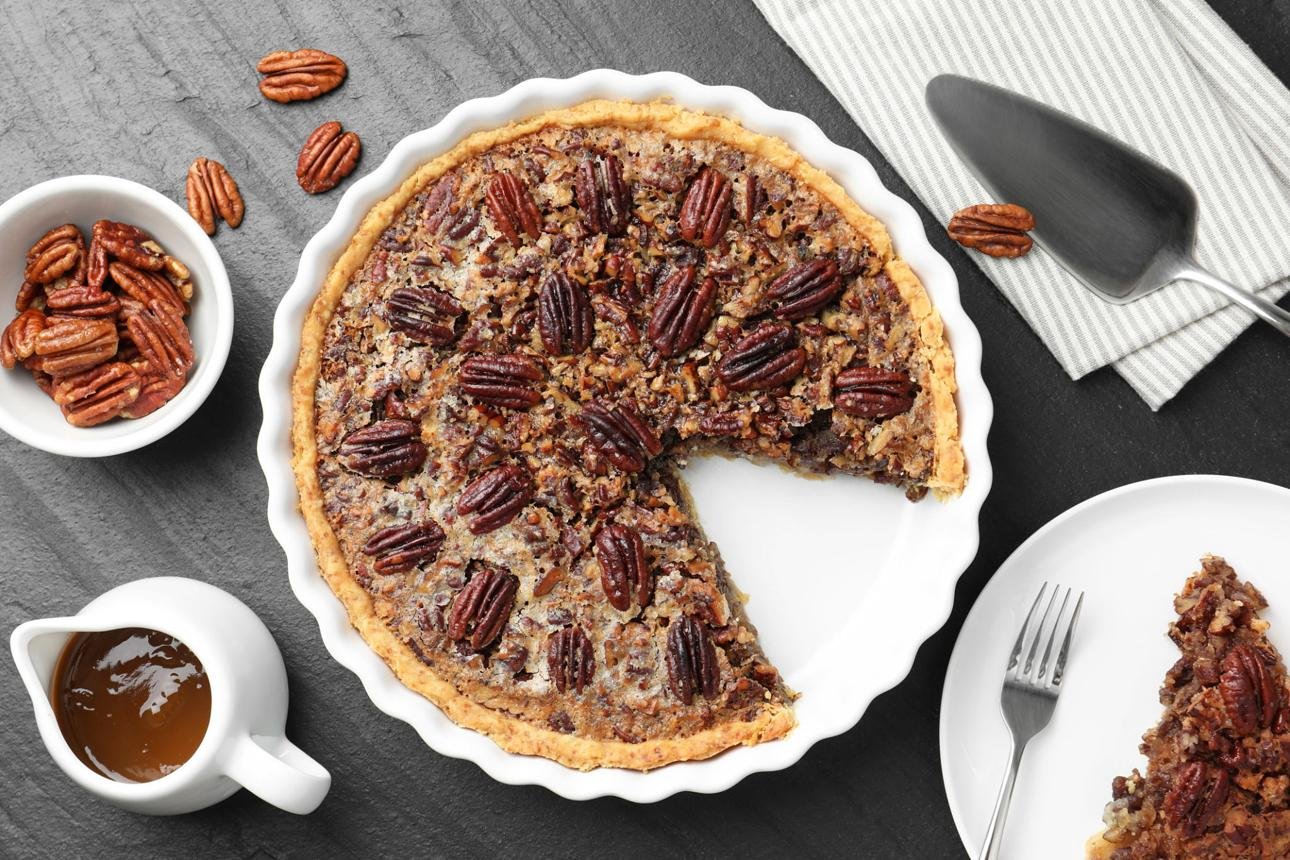 Pecan pie in a white fluted dish with a missing slice; pecans on top and a slice served on a plate to the right, with a bowl of pecans and caramel pitcher nearby.
