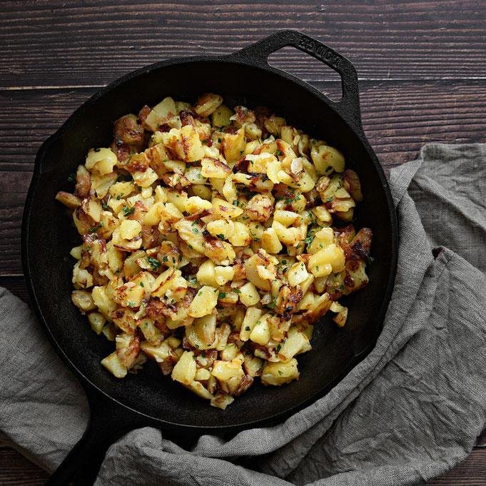 Skillet of crispy diced potatoes with herbs and bits of bacon, served in a cast-iron pan on a dark wooden table.