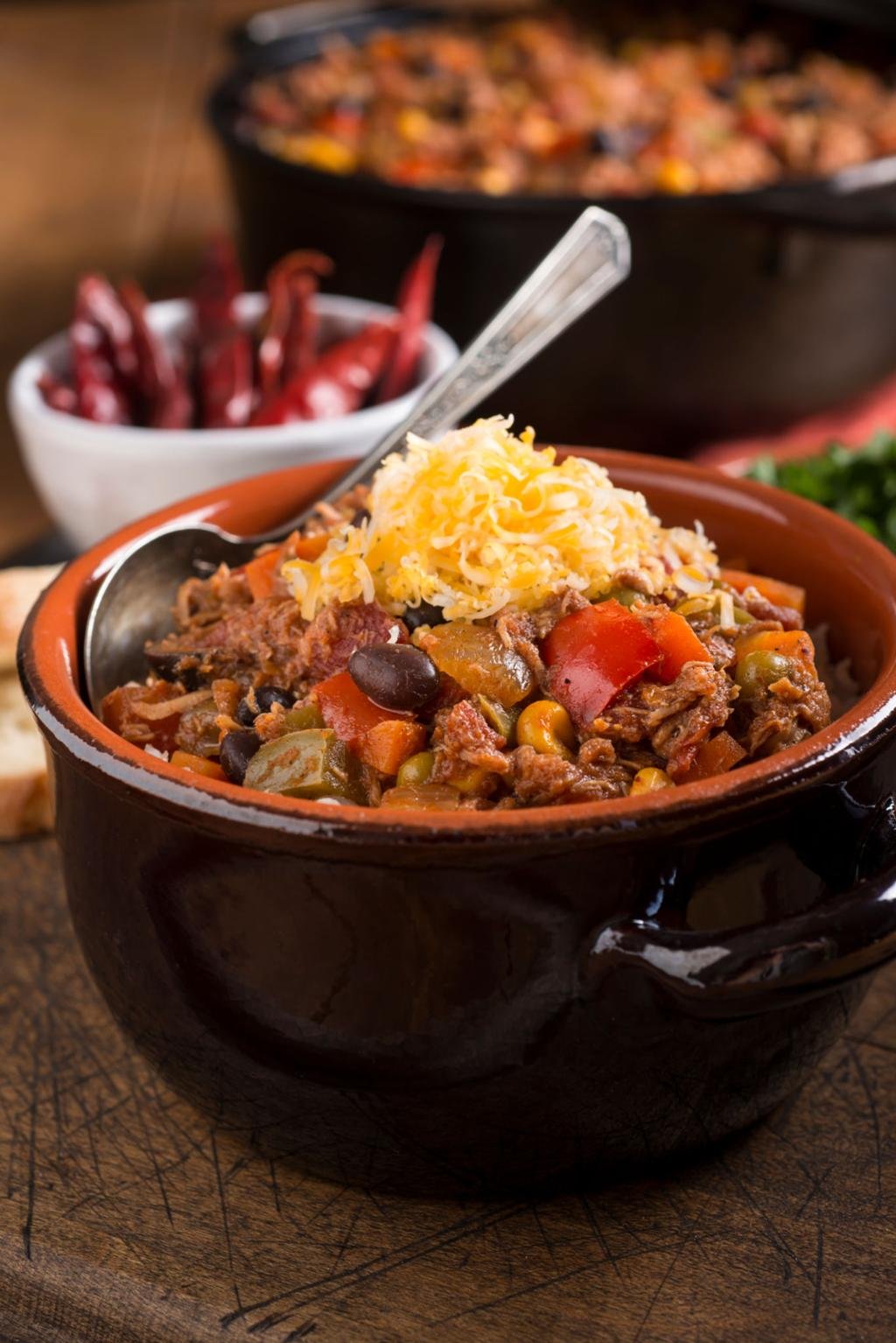 Beef chili in a brown ceramic pot topped with shredded cheddar, with beans and vegetables.