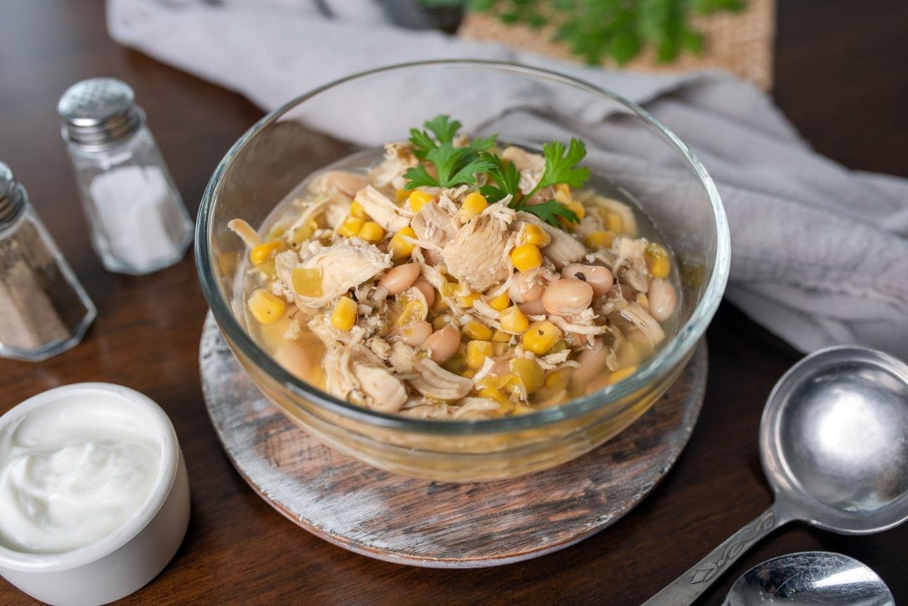 Glass bowl of chicken, corn, and white bean soup on a wooden board, garnished with parsley, with salt shakers and a spoon nearby.