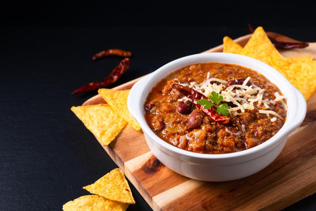 Bowl of chili topped with shredded cheese and cilantro on a wooden board with yellow tortilla chips nearby.