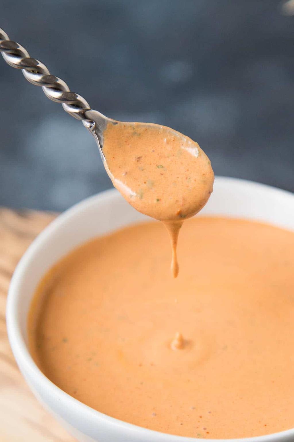 Creamy orange dipping sauce on a spoon, with a drip falling back into a white bowl below.