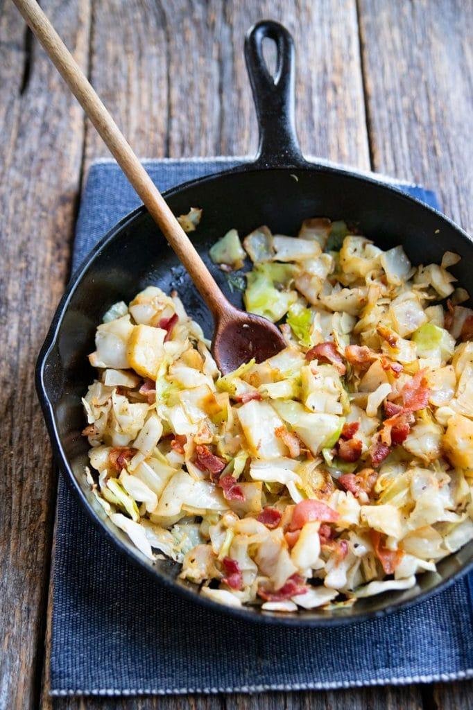 Cast-iron skillet on a wooden table, filled with cabbage and potato hash with bacon, wooden spoon resting inside.