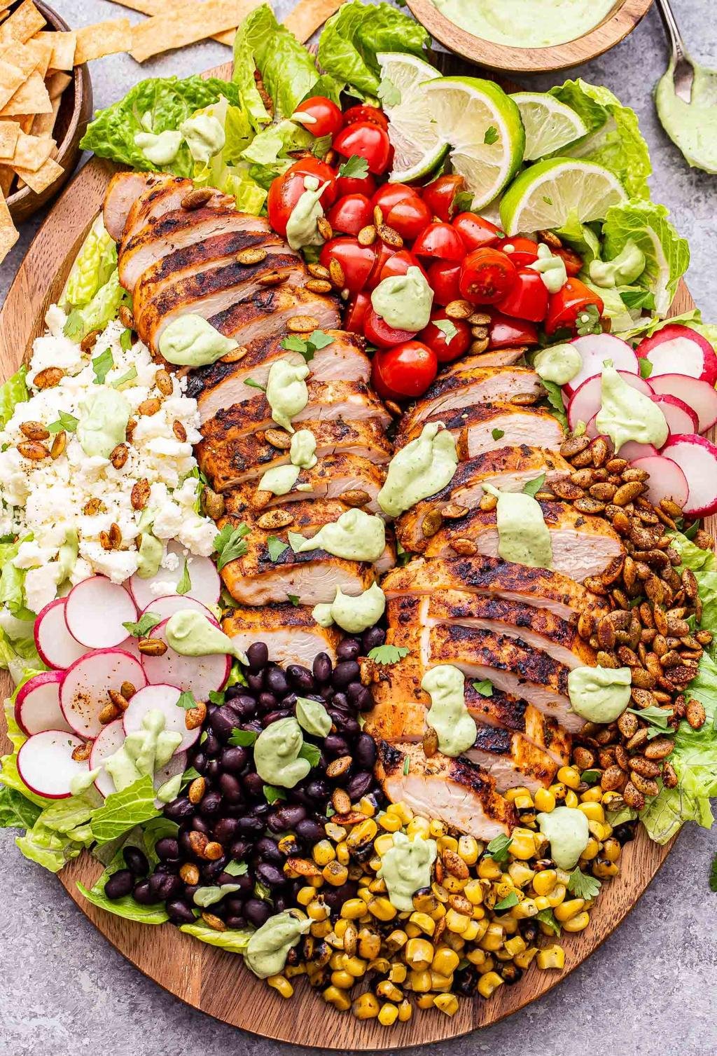 Sliced grilled chicken on a vibrant salad platter with tomatoes, radishes, corn, black beans, feta, avocado crema, and lime wedges.