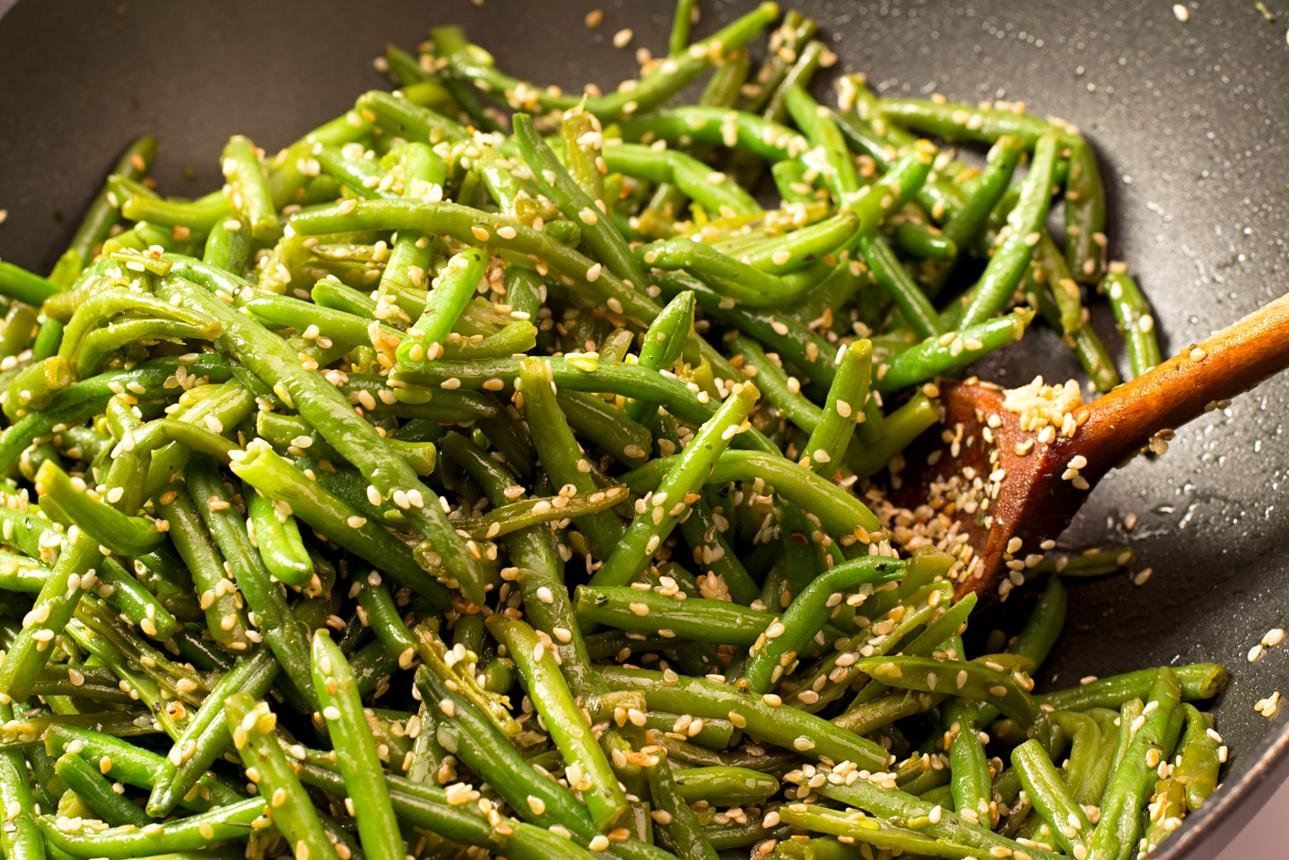 Stir-fried green beans with sesame seeds in a skillet, a wooden spatula resting nearby.