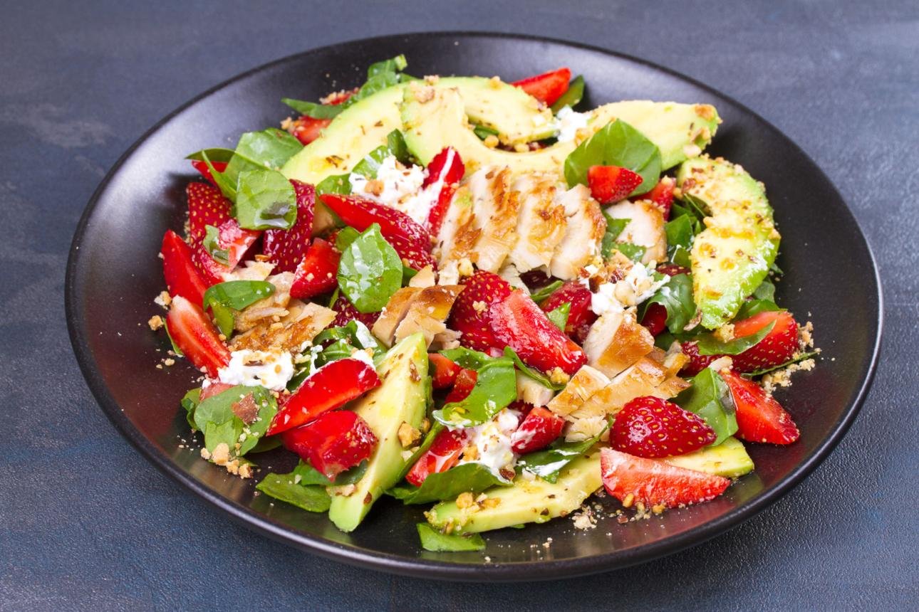 Plate of strawberry–avocado chicken salad with greens, feta, and chopped nuts on a dark plate.