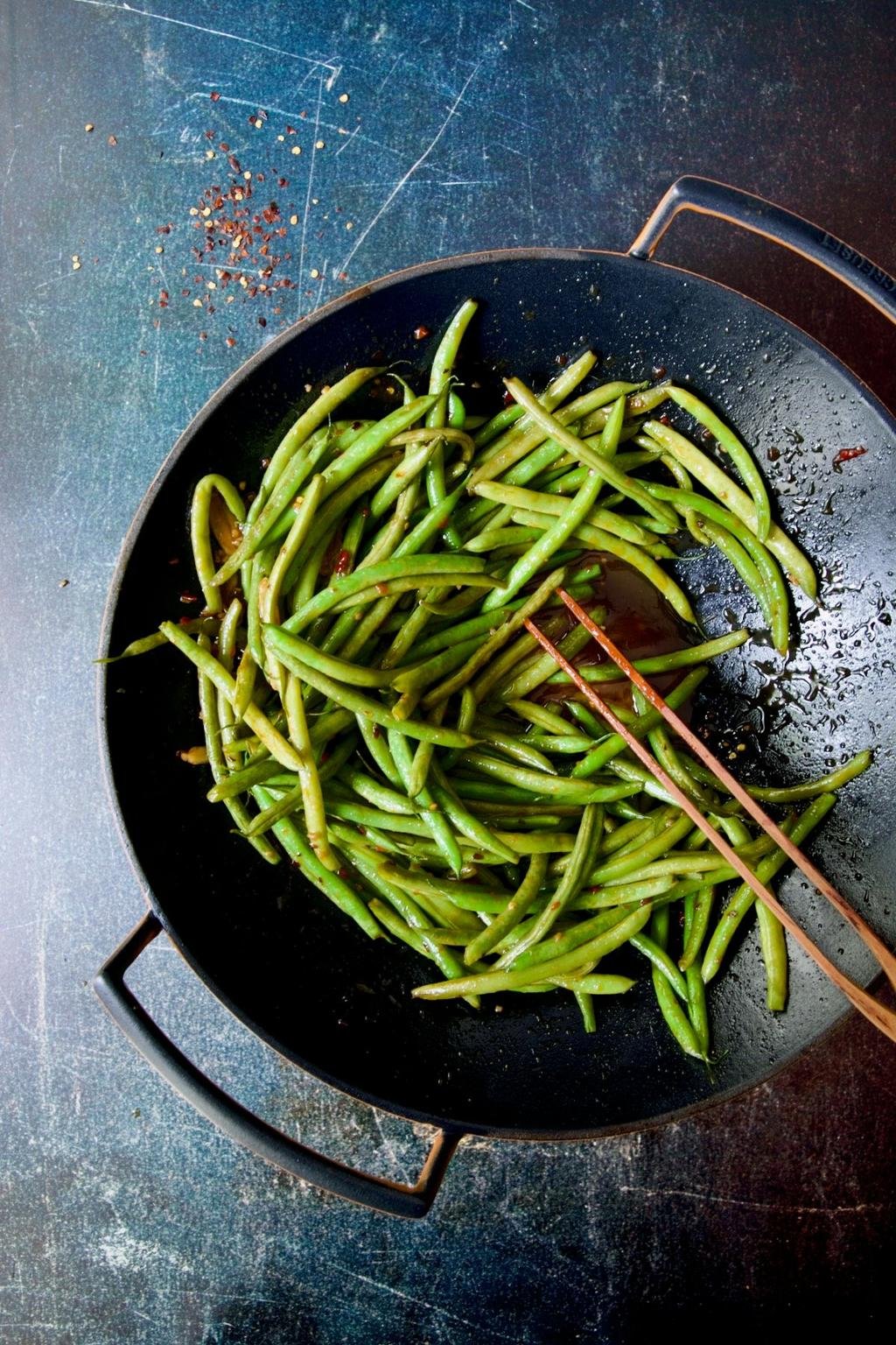 Sautéed green beans in a black skillet with chopsticks resting on the side, sprinkled with red pepper flakes.